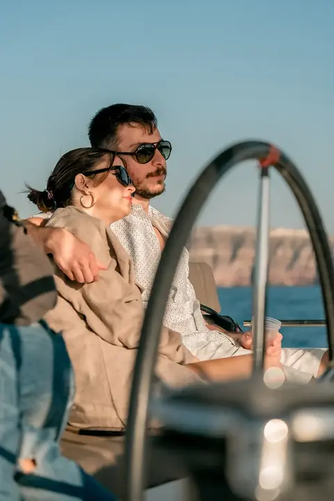 A couple seated together behind the catamaran's wheel in evening light, the caldera coast behind