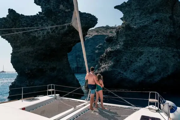 Two guests on the catamaran bow below the dark rock of the Akrotiri coast