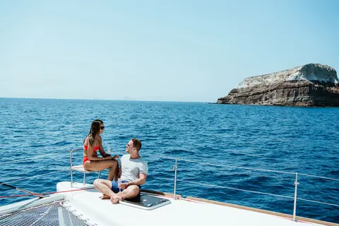 Two guests seated on the bow net of the catamaran off the pale cliffs of Santorini's south coast