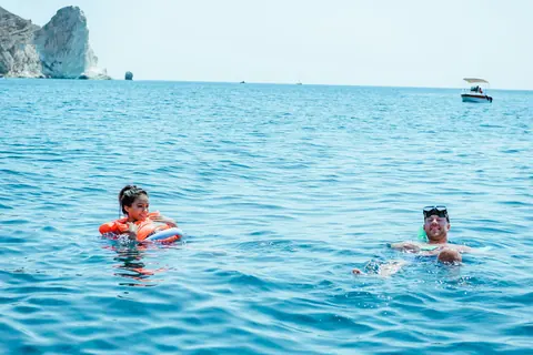Two swimmers with a float in open blue water, a pale rock headland in the background