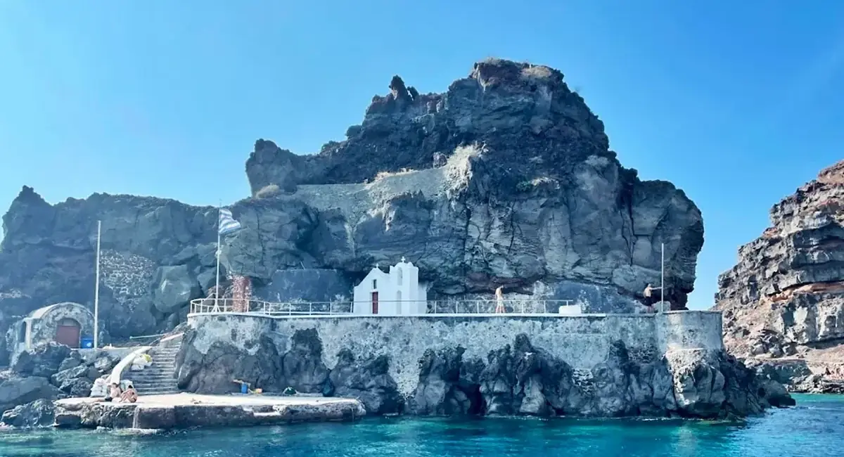 The white chapel of St. Nicholas on its rocky islet off Oia, with a Greek flag and a small landing stage
