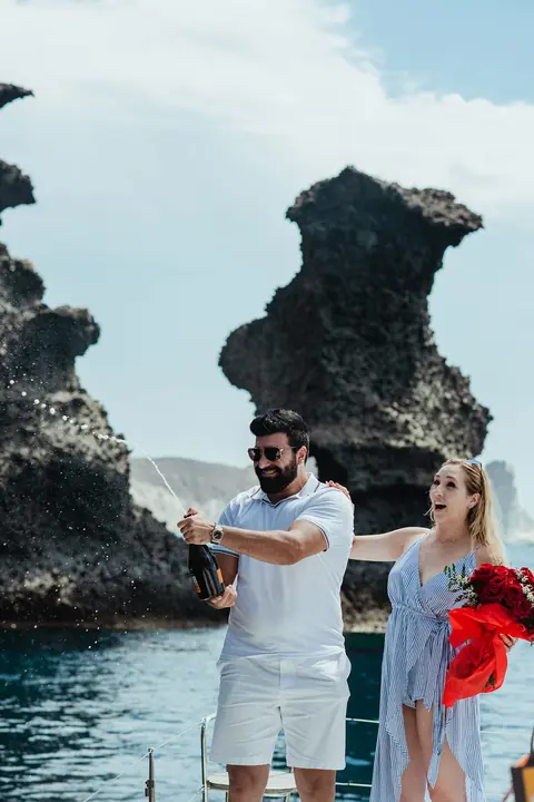 A couple opening champagne on deck with a bouquet of red roses, a volcanic sea stack behind
