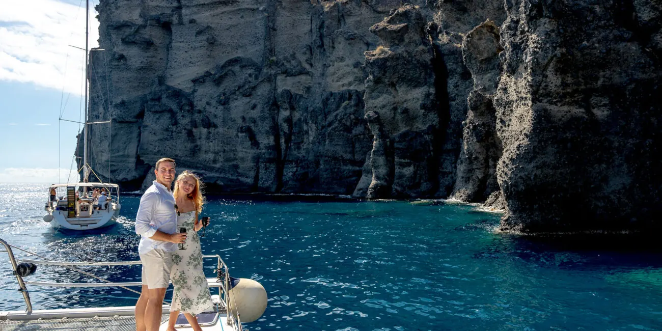 A couple with glasses on the bow of a catamaran below the pale cliffs of Santorini, a sailing yacht with guests alongside