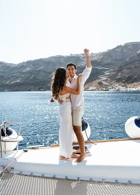 A couple celebrating on the bow of the catamaran with the zigzag path of Fira's old port behind