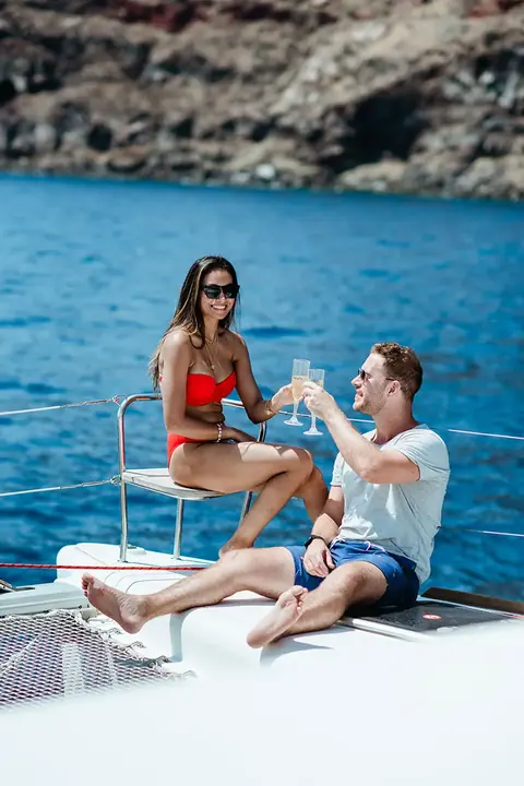 A couple raising glasses on the bow seat of the catamaran below the caldera cliffs