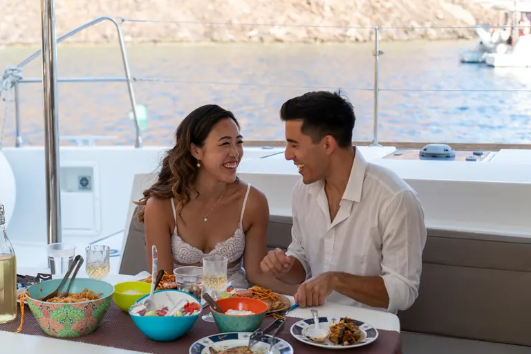 Two guests at the cockpit table of the catamaran with bowls of salad, pasta and grilled food, moored boats behind