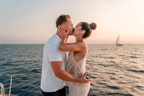 A couple kissing on deck at sunset with a sailboat on the horizon