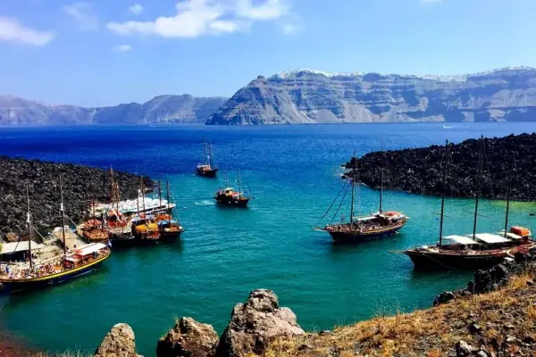 Wooden tour boats at the dark lava shore of the volcano, the caldera cliffs of Santorini across the water